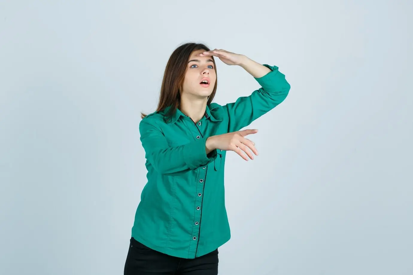 Junges Mädchen, das mit der Hand über dem Kopf in die Ferne blickt und von sich weg zeigt, in grüner Bluse und schwarzer Hose, wirkt konzentriert, Frontalansicht.